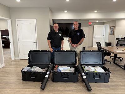 Two smiling men in black polo shirts stand in an office behind three open equipment cases filled with new school reunification equipment provided by BCOEM