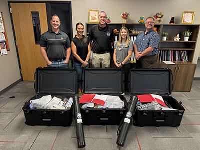Five adults pose behind three open black equipment cases filled with new school reunification equipment provided by BCOEM, creating a friendly team-presentation scene.
